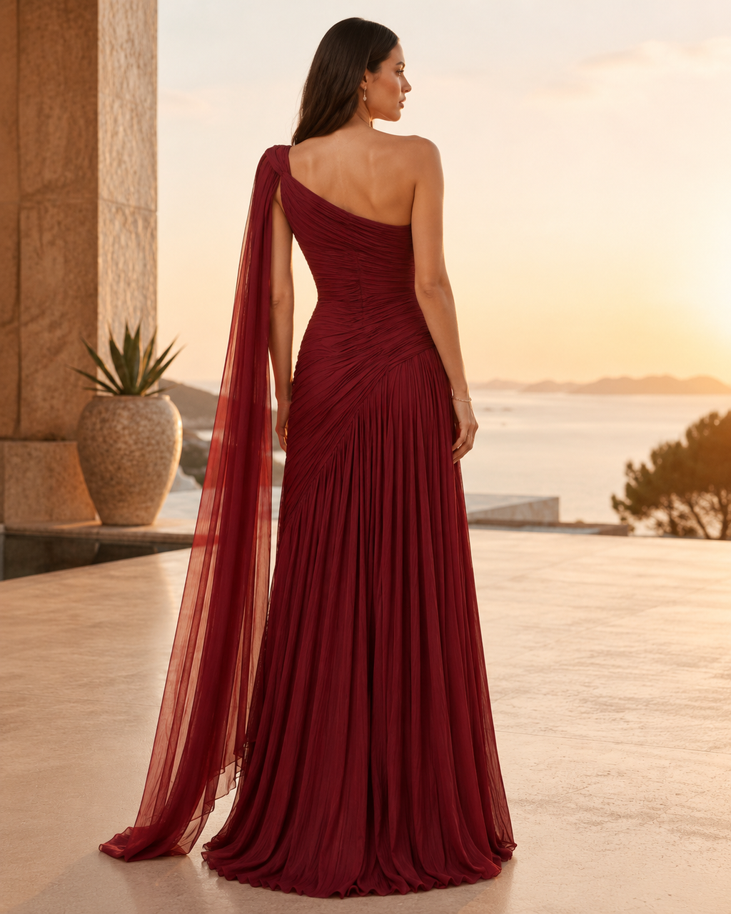 Wine one-shoulder evening gown shown from the back with pleated skirt and trailing sheer drape